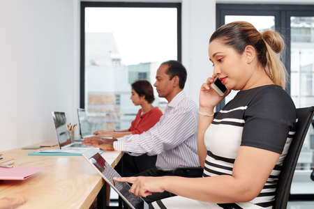 Positive Asian businesswoman reading document on tablet computer or browsing social media when calling on phone with clientの写真素材