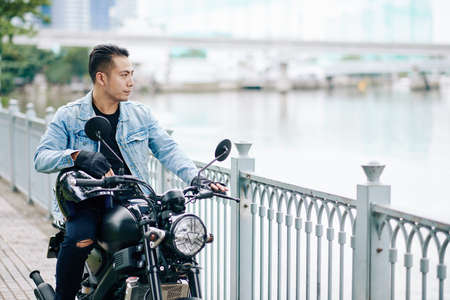 Handsome cool Asian man in denim jacket standing next to motorcycle and looking at riverの写真素材