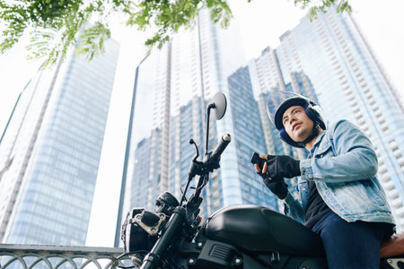 Handsome serious Vietnamese young man in helmet sitting on motorcycle with smartphone in hands,...