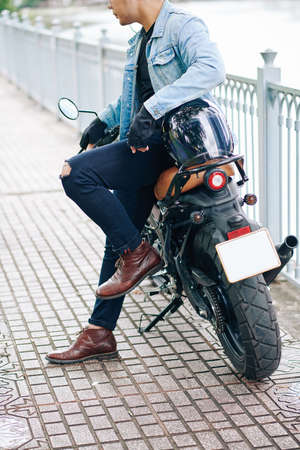 Cropped image of cool young man in leather boots and denim jacket leaning on his motorbike when standing outdoorsの写真素材