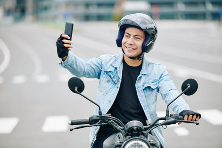 Happy handsome Vietnamese man in helmet taking selfie when sitting on his motorcycle