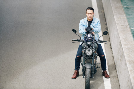 Serious attractive Vietnamese biker taking off helmet and looking at camera when sitting on...
