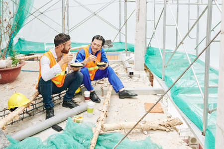 Positive engineer and contractor having lunch at construction site, eating noodles and discussing news and workの写真素材