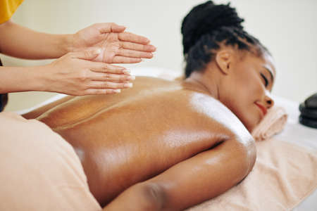 Close-up image of therapist massaging back of pretty young Black woman with organic oilsの写真素材