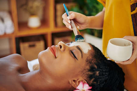 Close-up image of beautician applying purifying and minimizing pores clay mask on face of beautiful young Black womanの写真素材