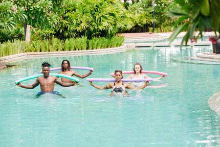 Multi-ethnic young people doing water aerobics with floating noodles in swimming pool of hotel
