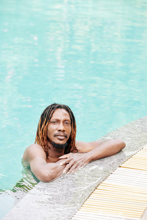 Portrait of serious handsome Black man with dreadlocks standing in swimming pool and looking at...