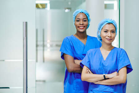 Smiling pretty multi-ethnic medical nurses in scrubs and disposable caps standing in hospital corridorの写真素材