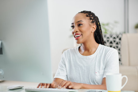 Cheerful young Black businesswoman sitting at office desk and answering e-mails from colleagues and...