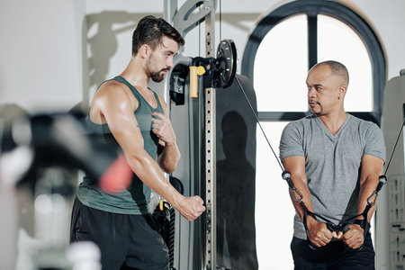 Fitness trainer explaining client what muscles should work when he is doing exercise in cable...