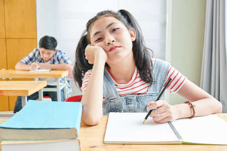 Bored Vietnamese schoolgirl sitting at desk in class and writing in copybookの写真素材