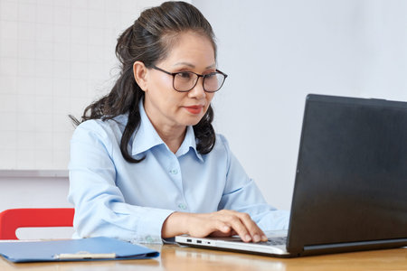 Mature Asian school teacher in glasses working on laptop at her table in class