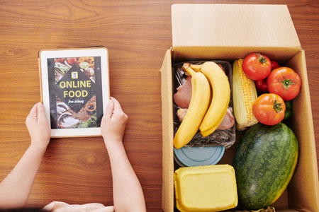 Housewife checking online food delivery application on tablet computer and unpacking big box of...