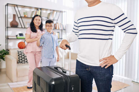 Cropped image of man standing in living room with big suitcase, his wife and teenage son waving in backgroundの写真素材