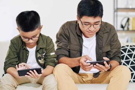 Vietnamese father and son in glasses sitting on sofa and plying games on smartphones