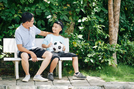 Vietnamese father and his preteen son resting on bench after playing soccer outdoors on Sundayの写真素材