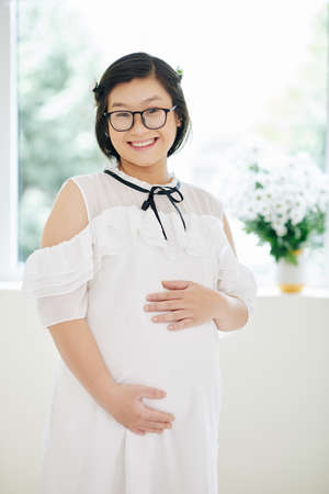 Portrait of smiling pregnant young woman in glasses touching her belly and looking at cameraの写真素材