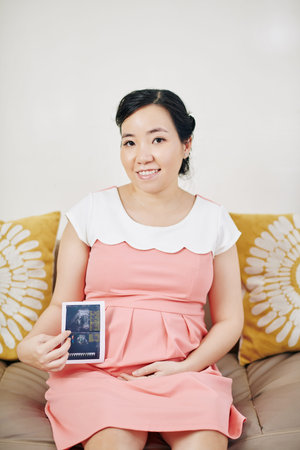 Portrait of smiling young pregnant Asian woman sitting on sofa and showing ultrasound scan of her...