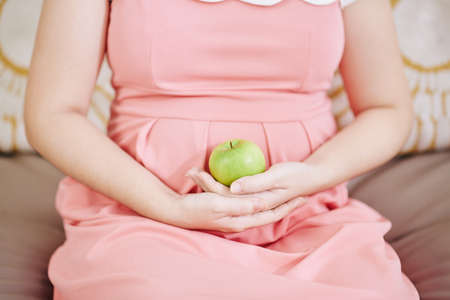 Cropped image of pregnant woman sitting on sofa with green apple in her handsの写真素材
