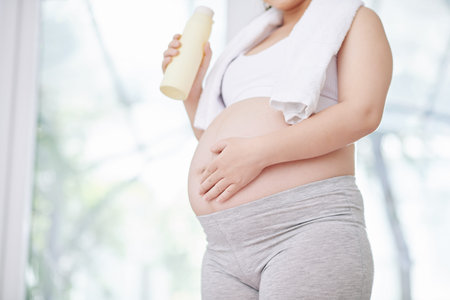 Cropped image of pregnant woman in crop top and leggings drinking water and touching her belly after...