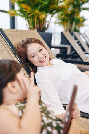 Happy Vietnamese mother and daughter resting on chaise-lounges outdoors and discussing newsの写真素材