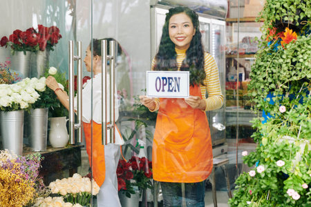 Beautiful young Vietnamese woman sticking open sign on glass door of flower shop