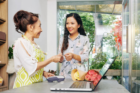 Smiling pretty young Vietnamese woman paying with credit card for bouquets she ordered in flower shopの写真素材