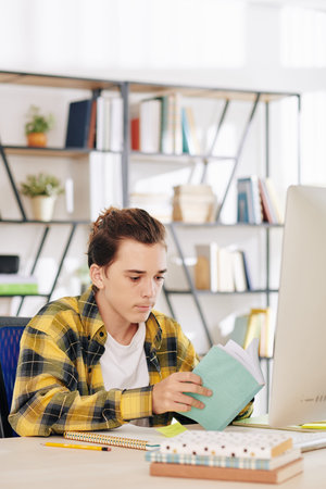 Serious teenage boy in black and yellow shirt sitting at desk at home and reading students books for...