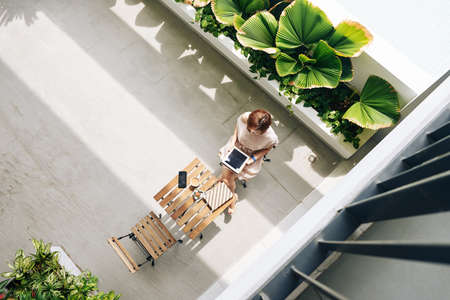 Young woman sitting at cafe table alone and reading article on digital tablet, view from aboveの写真素材