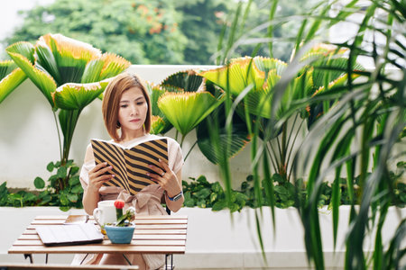 Serious pretty young Asian woman resting at cafe table and reading captivating bookの写真素材
