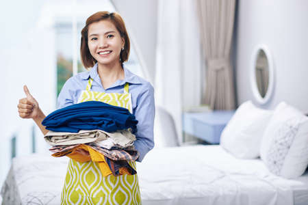 Portrait of smiling Vietnamese housewife holding pile of folded clothes she ironed and showing thumbs-upの写真素材