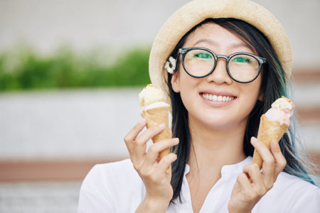 Portrait of attractive smiling young Chinese woman in glasses posing with delicious ice cream conesの写真素材