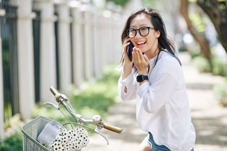 Portrait of happy laughing young Chinese woman sitting on bicycle and talking on phone with friend or boyfriendの写真素材