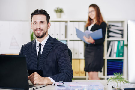 Portrait of cheerful young entrepreneur working on laptop at his office desk, his assistant...