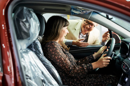Happy woman sitting in new car and satisfied her purchase with salesman standing outdoors and...