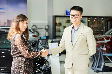 Portrait of Asian young manager in suit shaking hand to customer in car showroom
