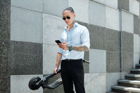 Serious frowning young businessman in glasses holding scooter and answering text messages from colleagues and clientsの写真素材