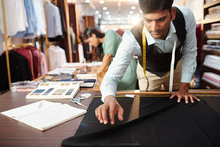 Professioal young Indian tailor laying out black fabric on big table in atelierの写真素材