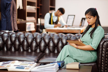 Beautiful young Indian woman sitting on sofa in atelier and watching catalog on tablet computer