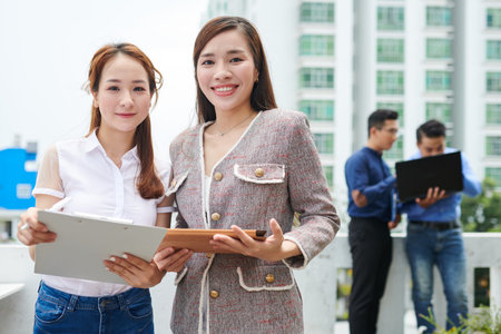 Portrait of young smiling Vietnamese businesswomen with clipboard and tablet computer standing...
