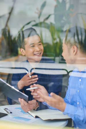 Cheerful yuong Asian entrepreneurs discussing plans of company development after analyzing reports at meetingの写真素材