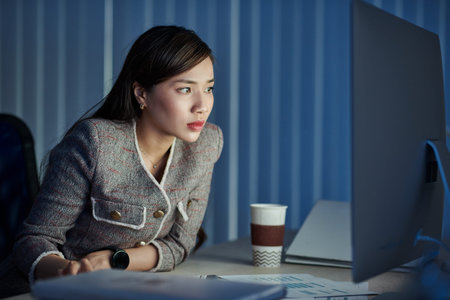 Serious young pretty business lady reading document on glowing screen when working in office late at...