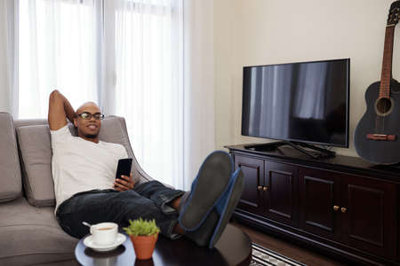 Lazy young Black man relaxing on sofa at home and checking text messages and social media on smartphoneの写真素材