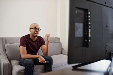 Excited young Black man watching sports competition on tv at homeの写真素材