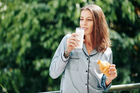 Beautiful young woman in pajamas having glass of milk or yogurt and croissant for breakfats when...