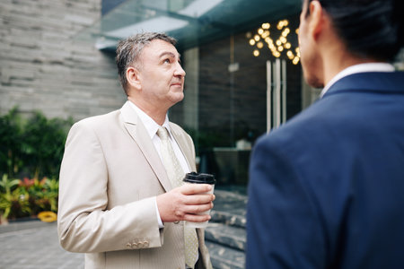 Pensive aged entrepreneur drinking morning coffee with his coworker and looking at office building...
