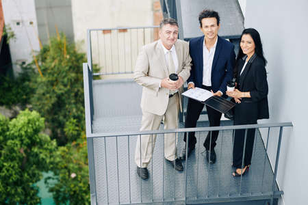 Smiling multi-ethnic business team with financial report standing on fire escape staircase and looking at cameraの写真素材