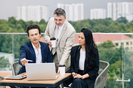 Serious business executives sitting at table at rooftop cafe and discussing data on laptop screenの写真素材