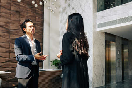 Serious mixed-race businessman talking to female entrepreneur in lobby of office buildingの写真素材