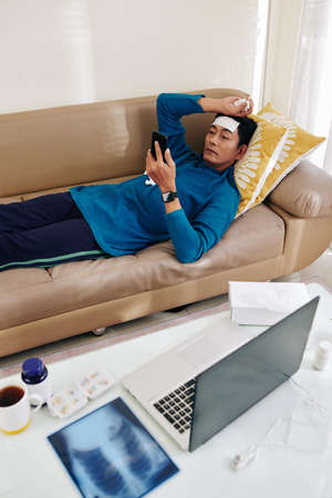 Man with cooling fever patch on his forehead lying on sofa and reading recommendations from his doctor on smartphoneの写真素材